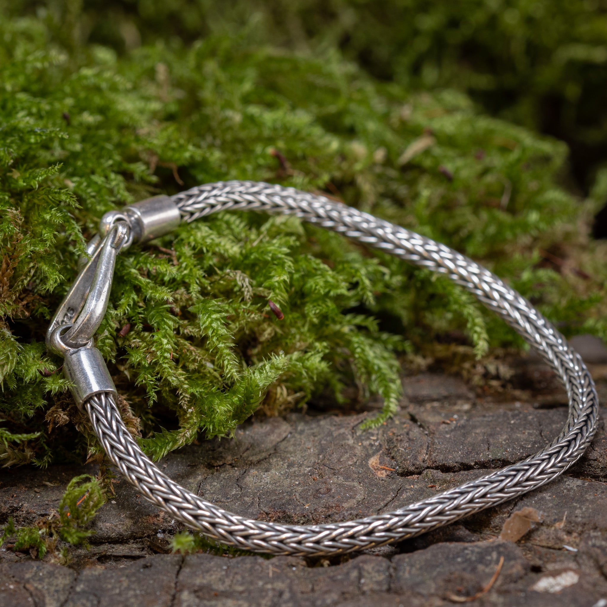 Et nærbillede af et Armbånd ByKila Sildeflet 3mm 925s Sølv flettet armbånd med en klassisk kroglås, placeret på en bund af grønt mos med et stykke bark under. Armbåndet står i kontrast til den naturlige baggrund og understreger dets indviklede design.