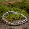 Load image into Gallery viewer, Et Armbånd ByKila Grov Rumpeflet 7mm 925s Sølv, komplet med sin indviklede lås, er elegant udstillet på en overflade af grønt mos og stykker af bark. Denne indstilling forstærker den naturlige og jordiske følelse af det polerede, flettet mønster smykke.
