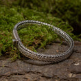 Load image into Gallery viewer, Et Armbånd ByKila Flad Sildeflet 4mm 925s Sølv med kasselås spænde ligger på en moset og stenet overflade. Armbåndet har et indviklet flettet-mønster, sat mod en naturlig baggrund af grønt mos og tekstureret bark.
