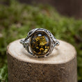 Load image into Gallery viewer, Et nærbillede af Sølvring Amber Envy 925s Sølv har et udsmykket sterlingsølvbånd og et rav ædelsten i midten, der viser indviklede blomsterlignende indeslutninger i rav. Fingerringen med grøn rav hviler på en tekstureret træoverflade med en sløret grøn løvbaggrund.
