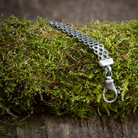 Et Armbånd Bismark 1,7 mm 925s Sølv med en hummerlås er draperet over en seng af levende grønt mos på en rustik træoverflade, der viser elegancen og kvaliteten af 925s Sterling sølv.