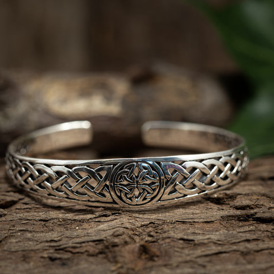 En Armbånd Bangle Crest 925s Sterling Sølv hviler på en træoverflade, med indviklede keltisk knude-design langs dens yderside med et centralt cirkulært motiv. Det justerbare armbånd har en let sløret baggrund, der afslører antydninger af grønt løv.