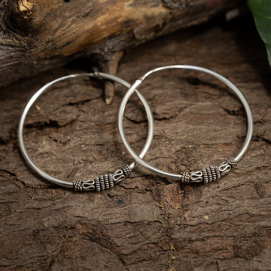 A pair of 925s silver hoop earrings rests on a textured wooden surface. The earrings have detailed decorative elements near the clasp, adding an artistic touch to the sleek circular hoops. Green foliage peeks in from the top right corner.