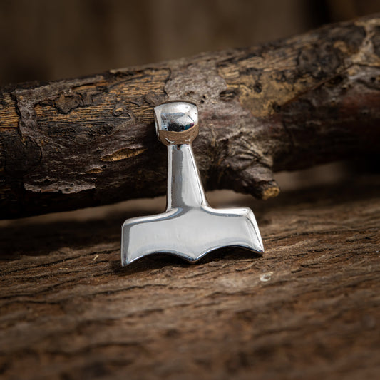 Et nærbillede af Thors Hammer Blanc Vedhæng 925s Sterling Sølv hvilende mod en forvitret træoverflade. Vedhænget har et simpelt design med et karakteristisk kort håndtag og et bredt, fladt hoved. Baggrunden inkluderer en træstamme med ru tekstur.