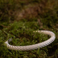 Load image into Gallery viewer, Et Armbånd Armbånd Empor 925s Sterling Sølv, et fantastisk sølvflettet armbånd, ligger på en seng af levende grønt mos. Det indviklede design står i kontrast til den naturlige, frodige baggrund, hvilket understreger dets detaljerede håndværk.
