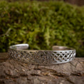 Load image into Gallery viewer, Armbånd Bangle Triskele 925s Sterling Sølv-armbånd hviler på en struktureret overflade og viser indviklede keltiske knudedesigns og et centralt cirkulært triskele-motiv. Baggrunden af sløret grønt understreger armbåndets detaljerede håndværk.
