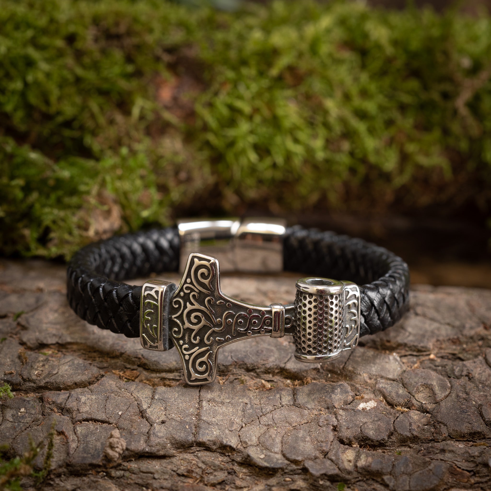 "Armbånd Thorshammer Flet m. Mønster Stål" er elegant udstillet på en rustik træflade med frodigt grønt mos i baggrunden. Det sorte læderarmbånd har en detaljeret sølv Thors hammer (Mjölnir) charme, og de indviklede designs på metaldelene skinner subtilt og tilføjer et nordisk twist mod den naturlige baggrund.