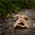 Load image into Gallery viewer, Broche Hugin og Munin Bronze, der er indviklet detaljeret for at fremhæve fjer og former for Odins ravne, hviler på tekstureret træbark med en plet af grønt mos i baggrunden.
