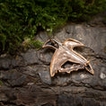 Load image into Gallery viewer, En lille, indviklet designet Broche Hugin og Munin Bronze i form af en fugl hviler på en struktureret træbarkoverflade med grønt og mos i baggrunden.
