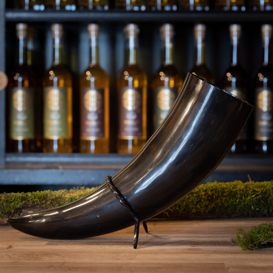 A polished black Drinking Horn Giant 1.5 liter rests on a stand before shelves of liquor bottles with gold circular logos. Moss covers the wood around it, adding a rustic touch reminiscent of Viking markets.