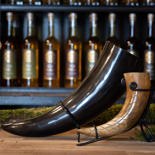 Two exquisitely crafted Drinking Horns, including Drinking Horn Giant 1.5 liters, in different sizes and colors, displayed on a wooden table. Each buffalo horn rests on a metal stand, with bottles visible on shelves in the rustic, elegant background, reminiscent of traditional Viking markets.