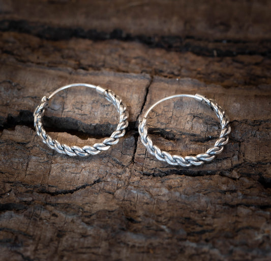 A pair of Braided 925s hoop earrings lies side by side on a textured wooden surface. Made of 925s Sterling Silver, their intricate braided design contrasts with the rustic background and highlights their delicate craftsmanship.