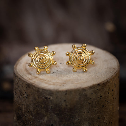 Close-up of Ear Studs Spiral Shield gold-plated 925s earrings, with concentric rings and small round bumps around the edge, made of gold-plated 925 Sterling Silver, resting on a rustic cylindrical wooden stand.