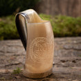 Load image into Gallery viewer, En Mini Vikingekrus Horn Thors Hammer (100 ml) sidder på en tekstureret stenoverflade, dens udsmykkede hornformede design minder om vikinge-drikkekar. Med et indgraveret cirkulært mønster med indviklede detaljer, muligvis forestillende et middelalder- eller fantasiemblem, kunne det nemt fremvises i Roskilde showroom. Baggrunden er sløret grønt og murværk.
