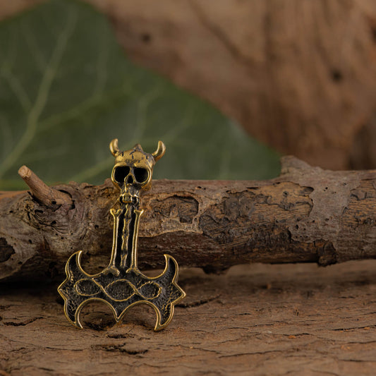 A brass Viking axe pendant with small horns and a black, intricate anchor-shaped body rests on a weathered tree branch, with green leaves in the background.
