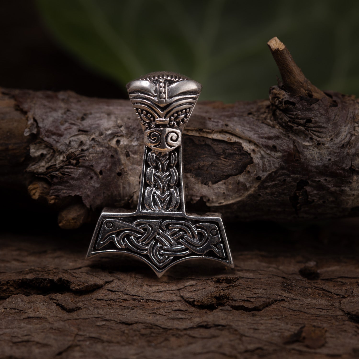 En detaljeret Thors Hammer Vedhæng 925s Sølv formet som Thors Hammer hviler på et groft stykke træ. Hammeren er lavet af 925s Sterling sølv og har indviklede nordiske designs og mønstre. Baggrunden er sløret med antydninger af mørkegrønt løv.