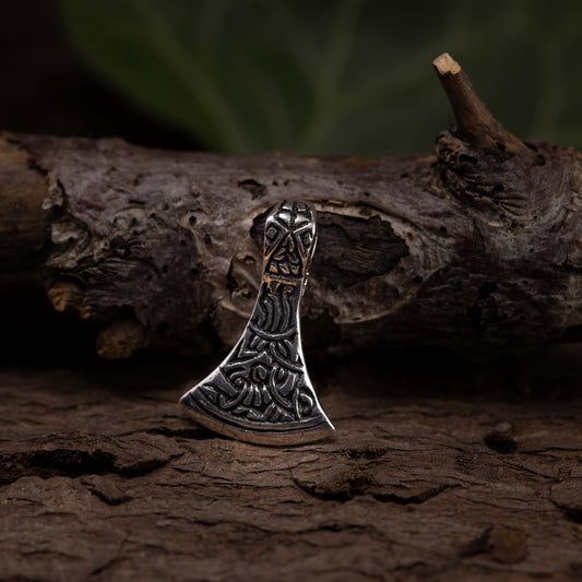 A small, intricately designed axe-head pendant in 925s silver rests on a textured wooden surface, with a blurred green background. The Viking jewelry has detailed engravings and appears inspired by Nordic or Viking motifs.