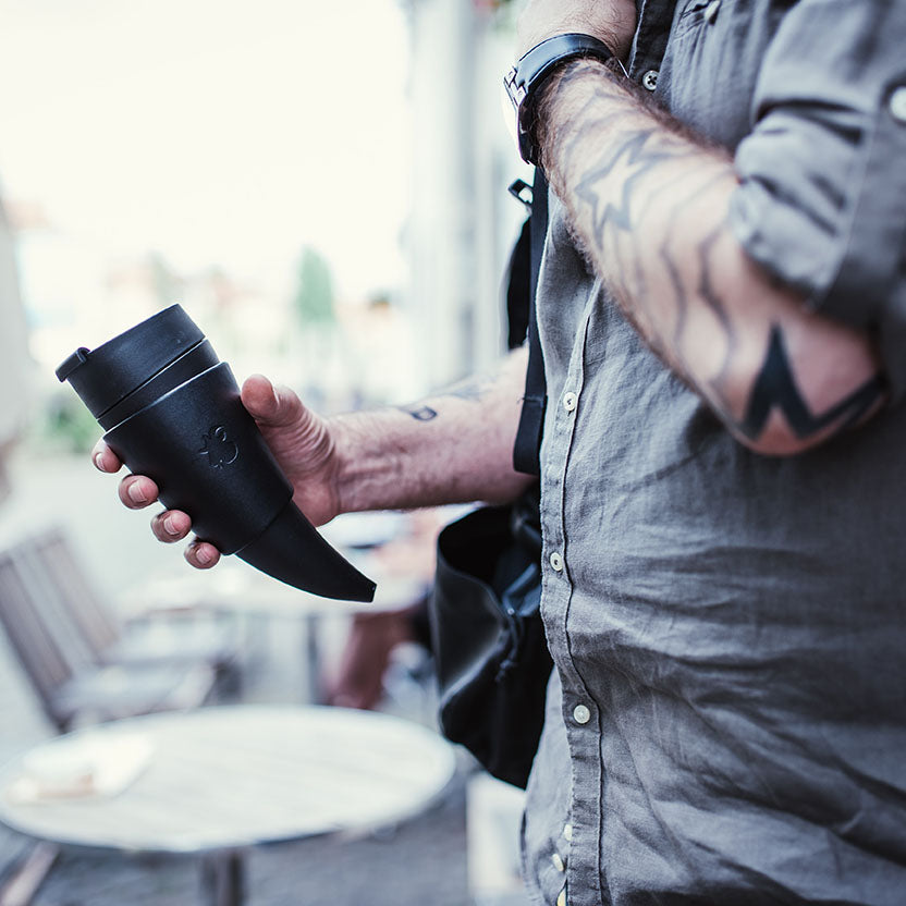 En person med tatoverede arme, iført en grå skjorte, holder en Goat Kaffe Drikkehorn Sort lavet af BPA-fri plast. De står udendørs nær et rundt bord med en sløret urban baggrund. En skuldertaskerem er synlig på deres højre skulder.