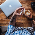 Load image into Gallery viewer, En person med tatoveringer på underarmen, iført en ternet skjorte, sidder ved et træbord. De holder en kop med latte art i den ene hånd og åbner låget på deres Ged Kaffe Drikkehorn Brun med den anden. En bærbar computer og et kamera er også på bordet, og fuldender scenen for denne moderne viking.

