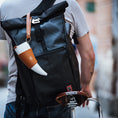 Load image into Gallery viewer, En person iført en grå t-shirt bærer en stor sort roll-top rygsæk med en hvid og solbrun ged Kaffe Drikkehorn Brun påsat, mens han skubber en cykel med en brun Brooks-sadel i læder. Scenen fremkalder stemningen fra en moderne viking på en bygade.
