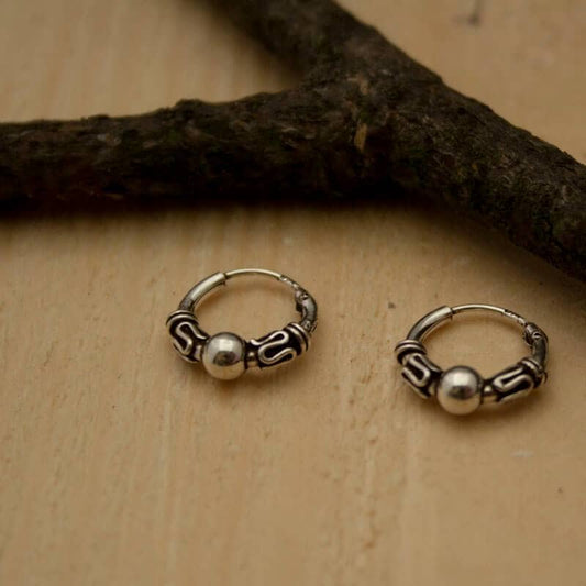 Wiuny 925s Silver Hoop Earrings, with intricate designs and a central silver bead, displayed on a light wooden surface. A dark, textured branch lies diagonally across the genuine silver earrings, inspired by medieval and Viking jewelry.