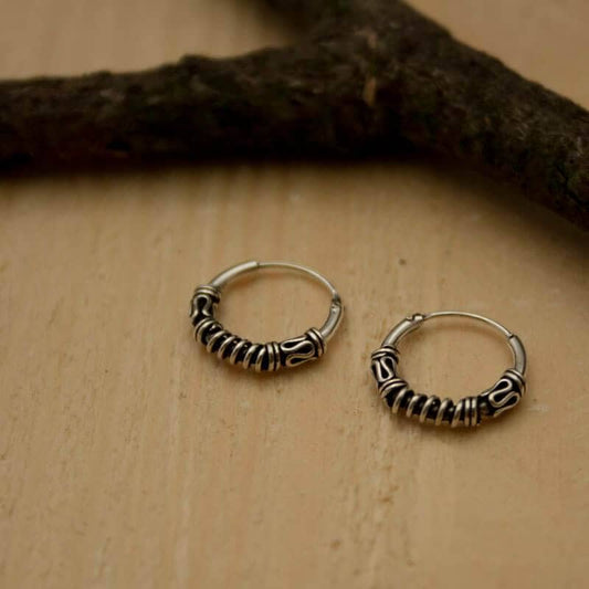 Two small, intricately designed silver hoop earrings with detailed bead patterns are placed on a light wooden surface. A dark, textured branch runs horizontally across the top of the image, adding rustic charm to these Olya 925s Silver Hoop Earrings.