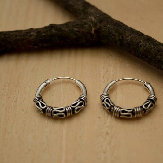 A pair of Avia 925s silver hoop earrings with intricate swirling designs rest on a light wooden surface. The earrings sit side by side near a dark branch placed diagonally across the upper left corner.