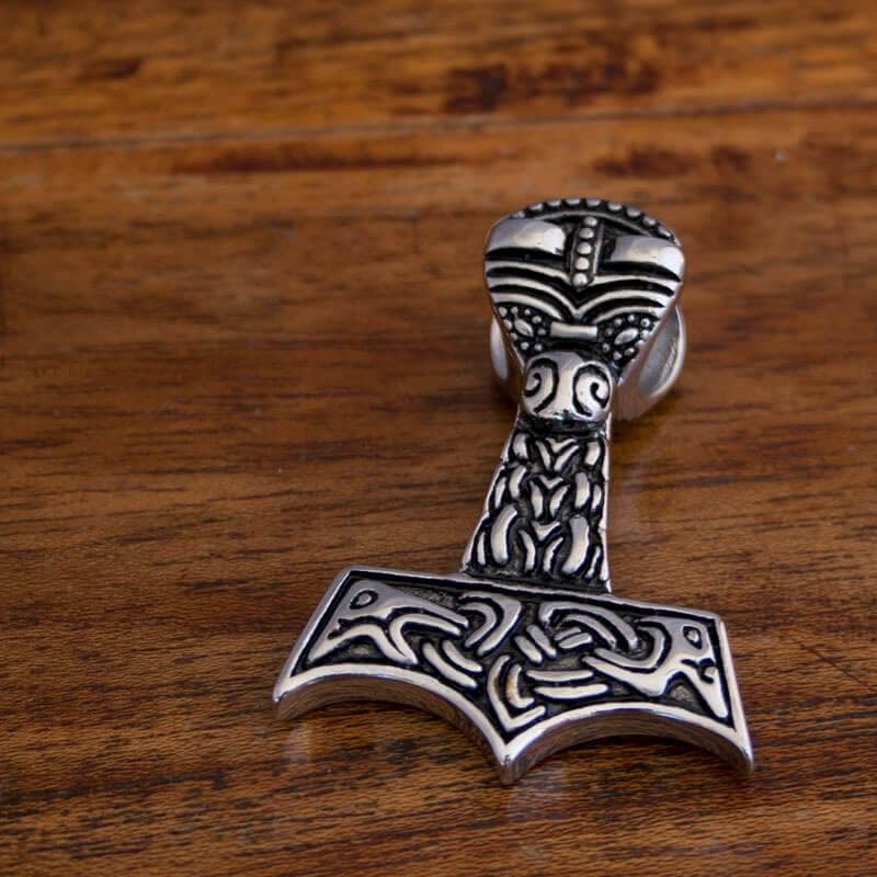 Et Thors Hammer Vedhæng Stål Anima vedhæng, formet som Mjölnir og prydet med indviklede nordiske designs, hviler på en træoverflade.