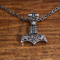 Load image into Gallery viewer, En sølvvedhæng med designet af Thors hammer, Mjolnir, med indviklede nordiske udskæringer og symboler på. Vedhænget er fastgjort til en Halskæde Ankerkæde Metallisk Stål Sort 5mm og hviler på en træoverflade.
