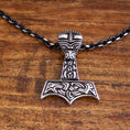 Load image into Gallery viewer, Et detaljeret vedhæng i form af Thors hammer, Mjölnir, hænger fra en Halskæde Ankerkæde Sort Stål 5mm. Vedhænget har indviklede, nordisk-inspirerede udskæringer og mønstre i en slående kontrastfarve. Den hviler på en træoverflade.
